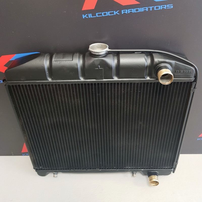 A classic car radiator finished in black, with a brass filler neck and outlet, against the KR backdrop